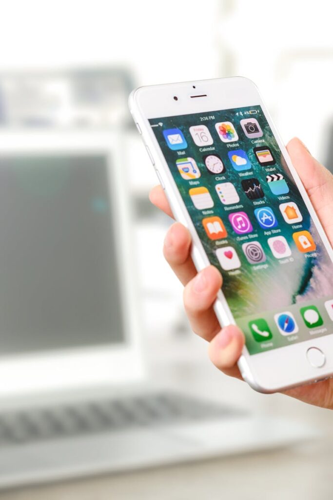 Close-up view of a smartphone displaying apps, held by a hand, with a blurred laptop in the background.